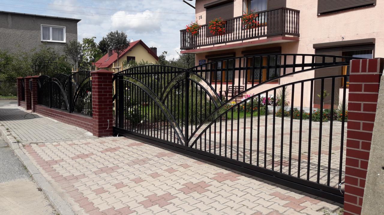 Czarna, przesuwna brama wjazdowa z metalowymi prętami i łukowym wzorem, osadzona na niskim murku z czerwonej cegły, na tle dwupiętrowego domu z balkonem ozdobionym czerwonymi kwiatami.