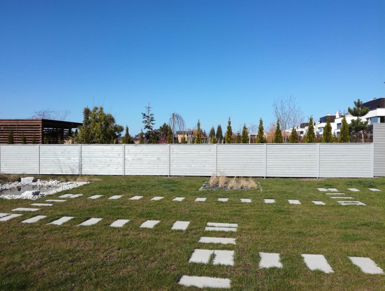 Długie, poziome ogrodzenie drewniane pomalowane na szaro, oddzielające trawnik z kamiennymi ścieżkami od drzew i budynków w tle, widoczne w słoneczny dzień.