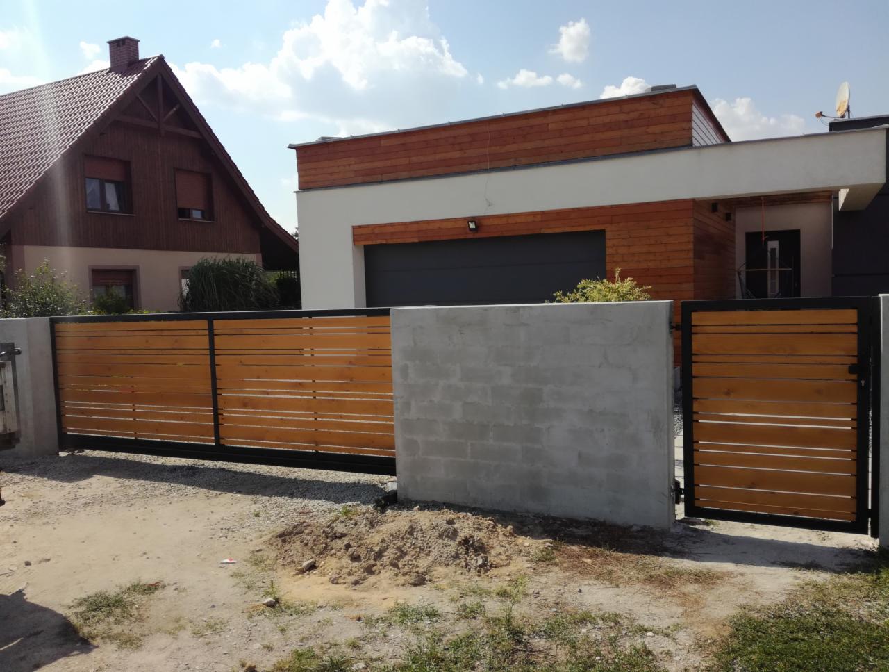 Suwana brama z poziomych desek w kolorze jasnego drewna, z czarną metalową ramą, pomiędzy betonowymi słupkami, na tle dwóch domów o różnej architekturze.