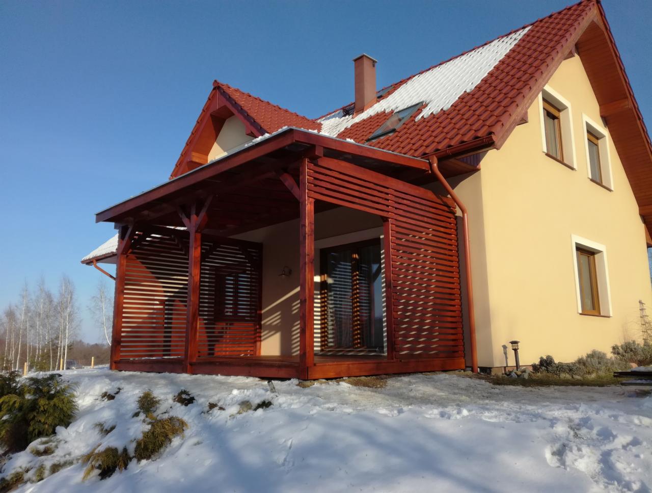 Dom jednorodzinny z dobudowanym, drewnianym zadaszeniem tarasu z poziomymi listwami, widok w zimowej scenerii, śnieg na dachu i ziemi, błękitne niebo.