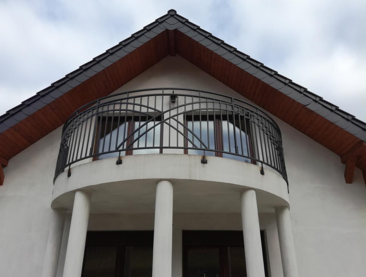 Elewacja budynku z balkonem o półokrągłej konstrukcji z kolumnami i czarną, kutą balustradą, widok z dołu na tle pochmurnego nieba.