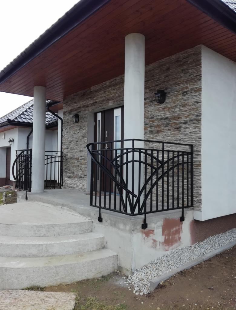 Czarna, metalowa balustrada z dekoracyjnym wzorem na betonowych schodach wejściowych do domu z elewacją z kamienia dekoracyjnego i białego tynku.