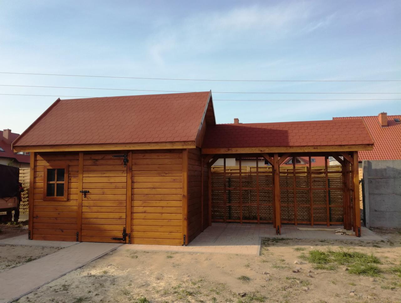 Drewniana konstrukcja z zamykaną częścią i zadaszeniem, wykończona czerwoną dachówką bitumiczną, z widocznym fragmentem posesji w tle.