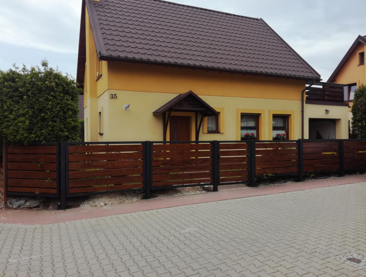 Nowoczesne ogrodzenie z poziomych desek w kolorze ciemnego drewna, zamontowane na czarnych, metalowych słupkach, przed żółtym domem z brązowym dachem.
