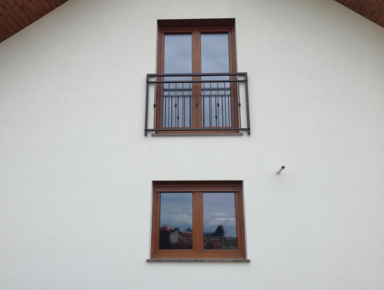 Elewacja domu z dwoma oknami; górne okno z czarną, metalową balustradą balkonową o prostym wzorze.