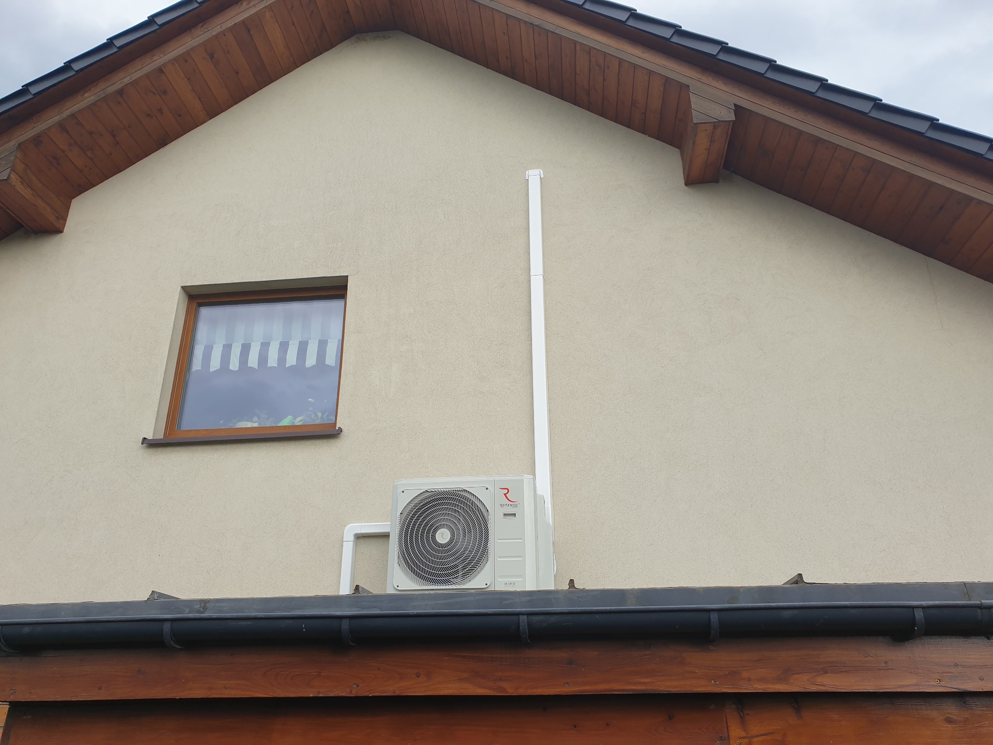 Zewnętrzna jednostka klimatyzacji Rotenso HIRO zamontowana na elewacji domu, widoczna rura odprowadzająca skropliny i okno z żaluzjami w tle.