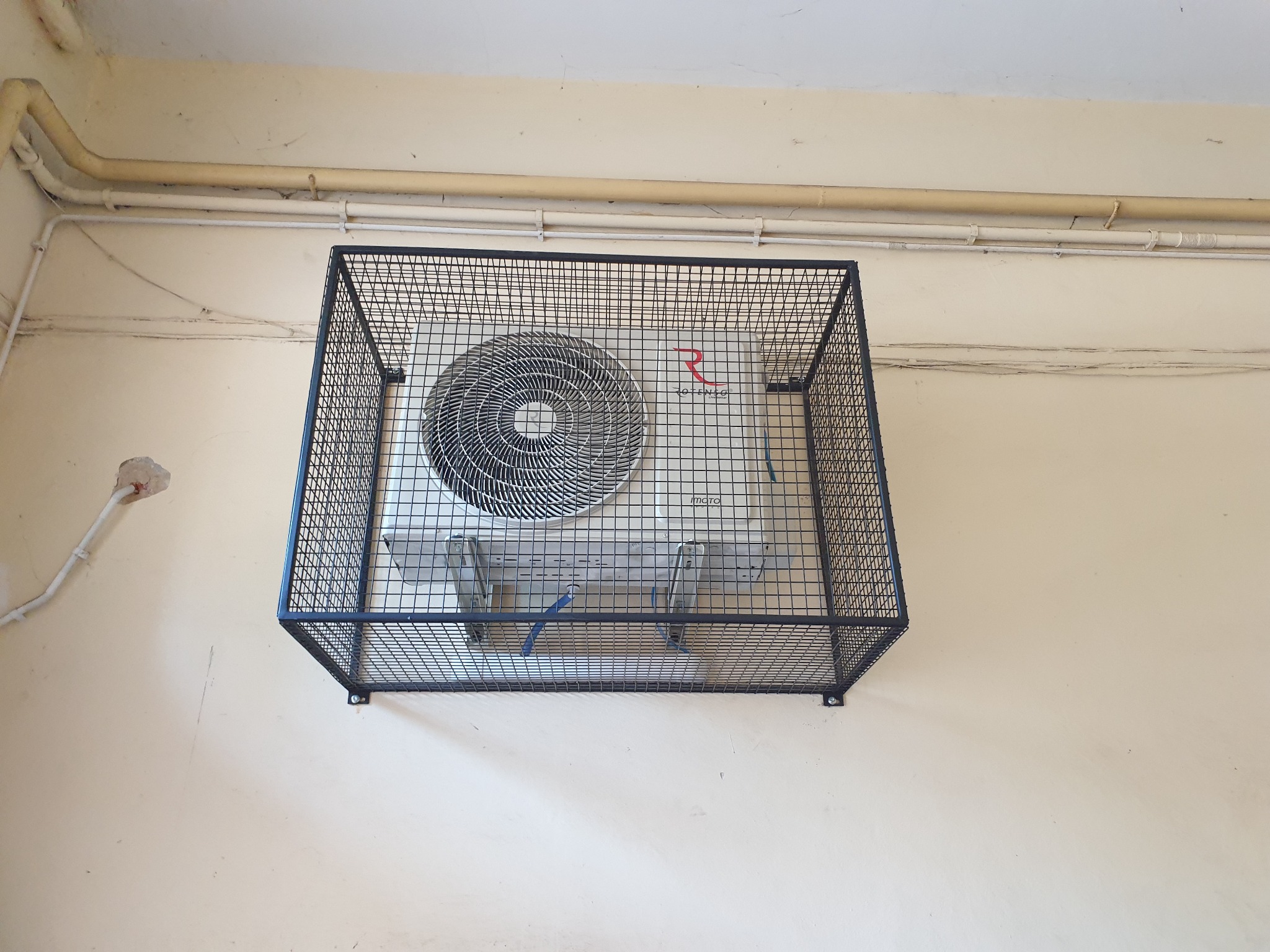 Zewnętrzna jednostka klimatyzacji marki Rotenso, zabezpieczona metalową klatką, zamontowana na ścianie w Jenkowicach. Widoczne rury instalacyjne pod sufitem.