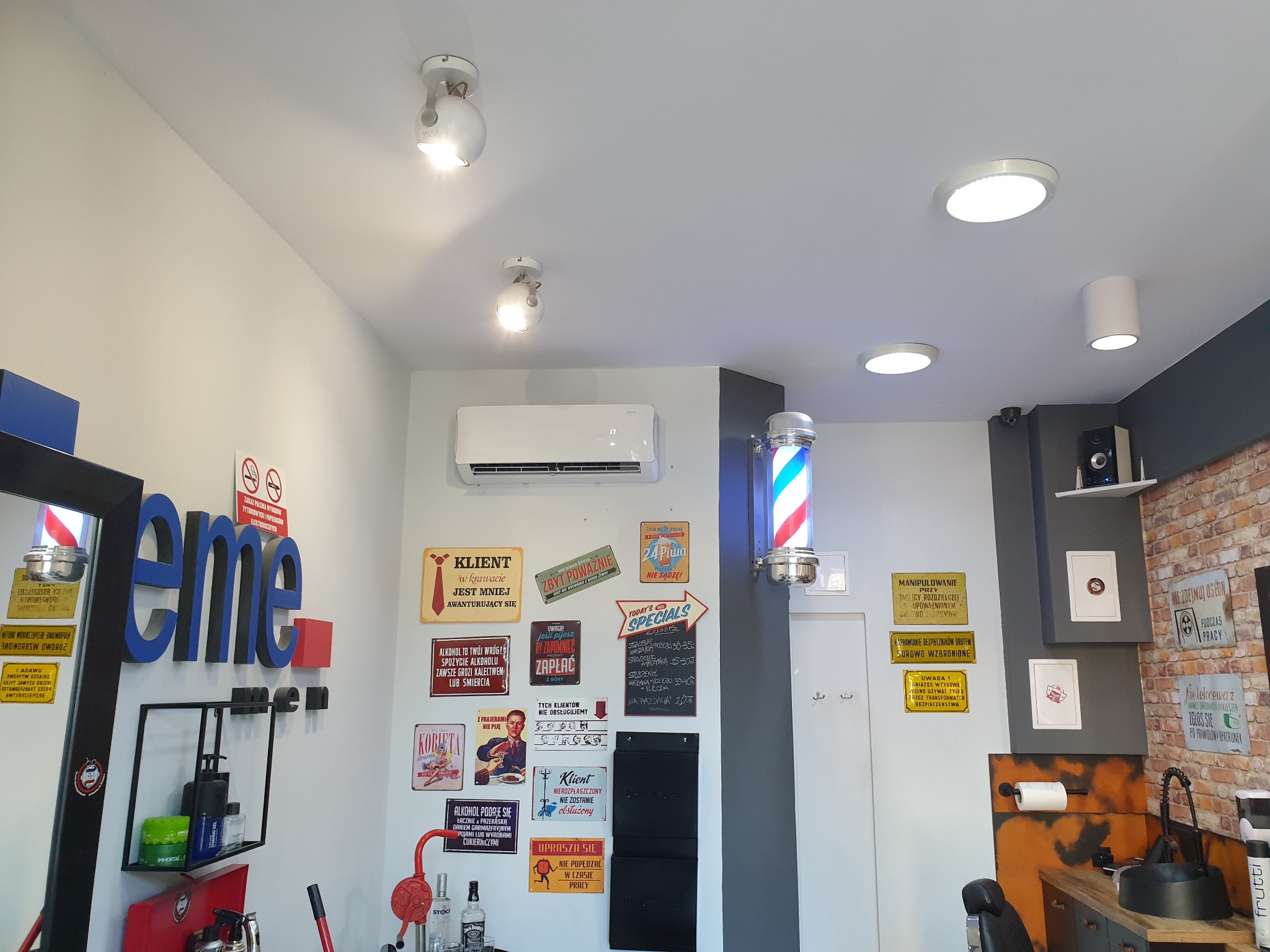 Wnętrze salonu fryzjerskiego z klimatyzacją ścienną, oświetleniem punktowym i dekoracyjnymi tabliczkami w stylu retro.