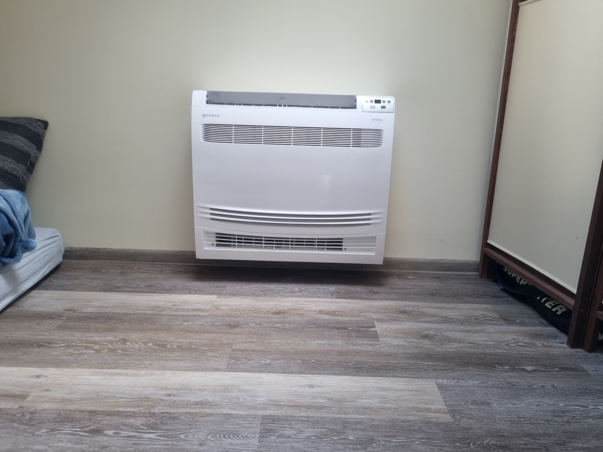 Biała, przypodłogowa jednostka klimatyzacyjna marki Rotenso, zamontowana na kremowej ścianie w pomieszczeniu z panelami podłogowymi o wzorze drewna, widoczny fragment sofy i ramy okiennej.