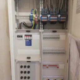 Otwarta skrzynka rozdzielcza z zamontowanymi bezpiecznikami, przewodami i listwami zaciskowymi, widoczne dokumenty wewnątrz obudowy.