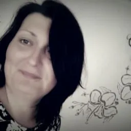 Uśmiechnięta kobieta z czarnymi włosami, w bluzce w kwiaty, na tle ściany w kwiatowe wzory. Portret w sepii.