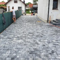 Ułożona kostka brukowa w odcieniach szarości na podjeździe, z widoczną zagęszczarką i pracownikiem opartym o zielone ogrodzenie, w tle domy jednorodzinne.