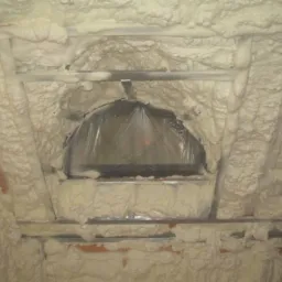 Poddasze z oknem dachowym, zabezpieczonym folią, pokryte grubą warstwą jasnej, spienionej izolacji natryskowej. Widoczne metalowe profile konstrukcyjne.