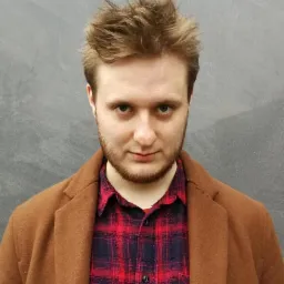 Portret młodego mężczyzny z rudą brodą, ubranego w brązowy płaszcz i czerwoną koszulę w kratę, na tle szarej ściany. Ujęcie od przodu, skupione na twarzy.