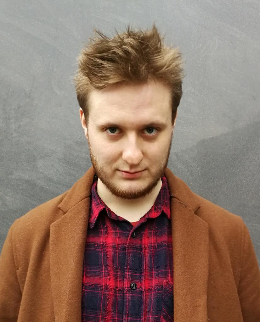 Portret młodego mężczyzny z rudą brodą, ubranego w brązowy płaszcz i czerwoną koszulę w kratę, na tle szarej ściany. Ujęcie od przodu, skupione na twarzy.