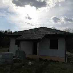 Dom w trakcie budowy z ciemnoszarą dachówką, widoczne materiały budowlane i fragment lasu w tle.