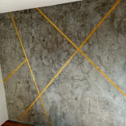 Szara ściana z efektem betonu architektonicznego, zdobiona złotymi, ukośnymi pasami tworzącymi geometryczny wzór.