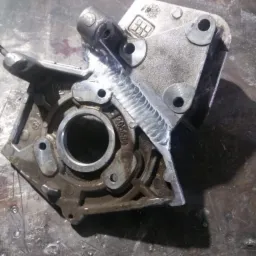 Szara, metalowa obudowa elementu maszynowego po spawaniu, widoczne spawy na krawędziach i wok&oacute;ł otworu, zabrudzenia na powierzchni, metalowy st&oacute;ł warsztatowy w tle.