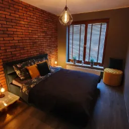 Sypialnia z łóżkiem z ciemną pościelą, ceglaną ścianą oświetloną ciepłym światłem, lampką nocną na stoliku i geometryczną lampą wiszącą.