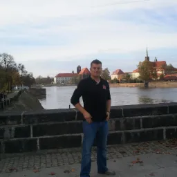 Mężczyzna w czarnej koszulce polo pozuje na tle Ostrowa Tumskiego we Wrocławiu, stojąc na kamiennym moście. Widoczne jesienne liście i fragment rzeki.