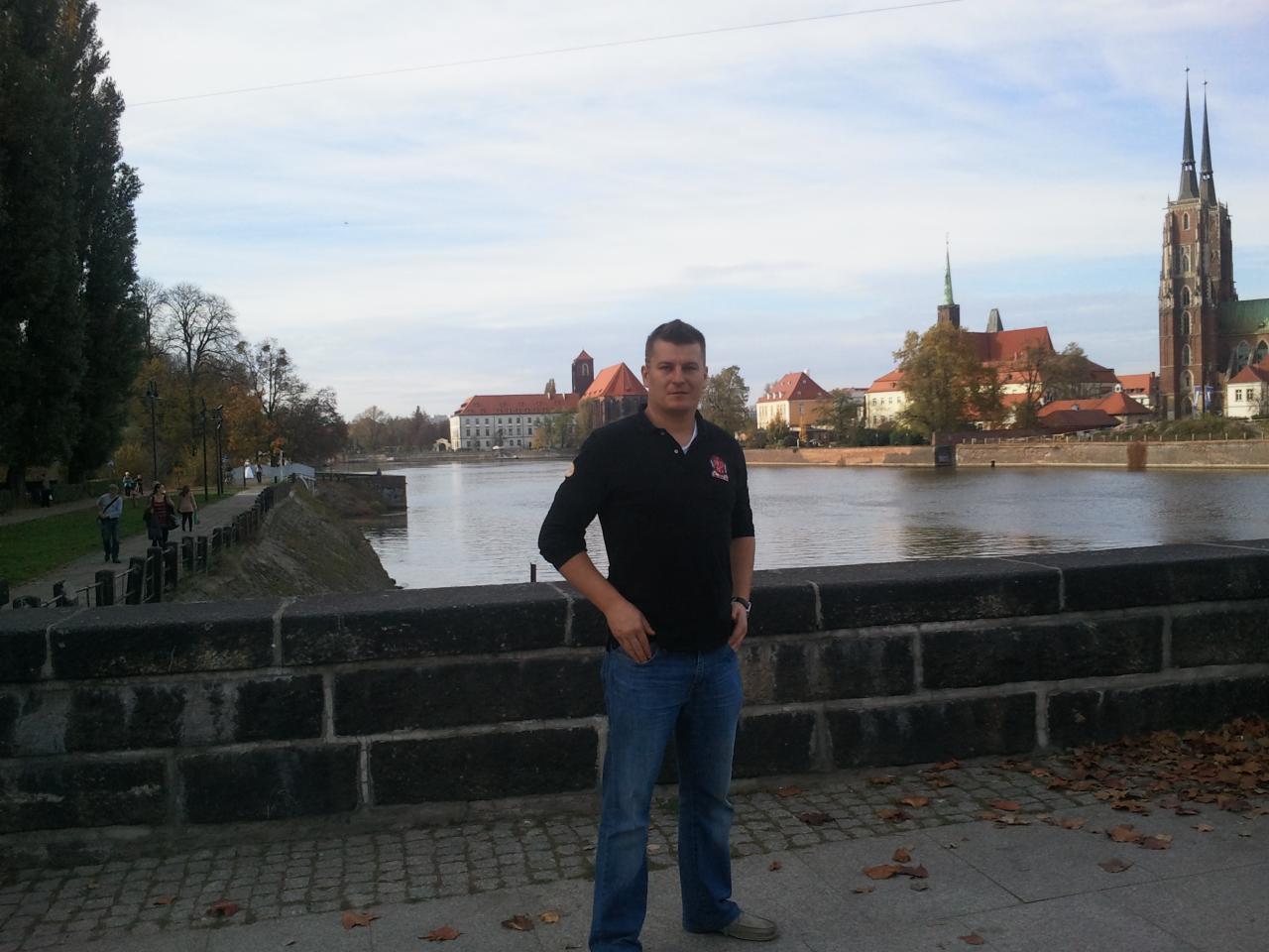 Mężczyzna w czarnej koszulce polo pozuje na tle Ostrowa Tumskiego we Wrocławiu, stojąc na kamiennym moście. Widoczne jesienne liście i fragment rzeki.