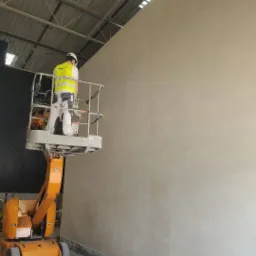 Pracownik w kasku i kamizelce odblaskowej na podnośniku koszowym maluje jasną ścianę w hali przemysłowej. Widoczna faktura świeżo nałożonej farby.