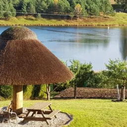 Altana kryta strzechą nad jeziorem, z drewnianymi stołami i ławkami, otoczona zielenią i drzewami iglastymi w tle.