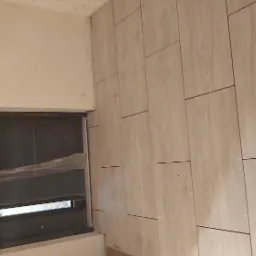 Ściana wykończona panelami imitującymi drewno, ułożonymi pionowo, z widocznym oknem zabezpieczonym folią ochronną podczas prac.