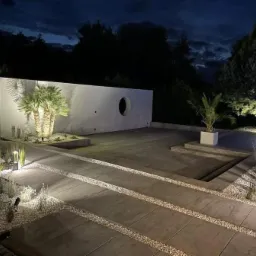 Nowoczesny ogród w Bieżeniu oświetlony nocą, z minimalistycznym murem z okrągłym oknem, palmami i ścieżką z płyt betonowych wysypaną białym żwirem.