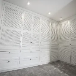 Biała, zabudowana szafa z lamelowymi drzwiami i szufladami, oświetlona punktowymi lampami sufitowymi, na szarym dywanie.