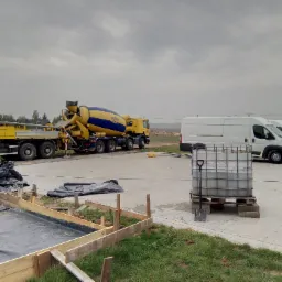 Przygotowanie pod wylewkę: szalunek z desek, folia izolacyjna, obok betoniarka i dwa busy na placu budowy w pochmurny dzień.