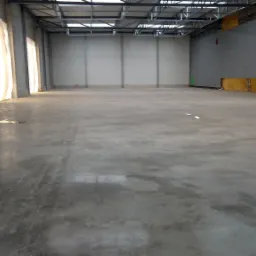 Betonowa posadzka w hali przemysłowej przed oddaniem do użytku, widoczne zabezpieczenia ścian i sufitu, naturalne światło wpadające przez bramę.