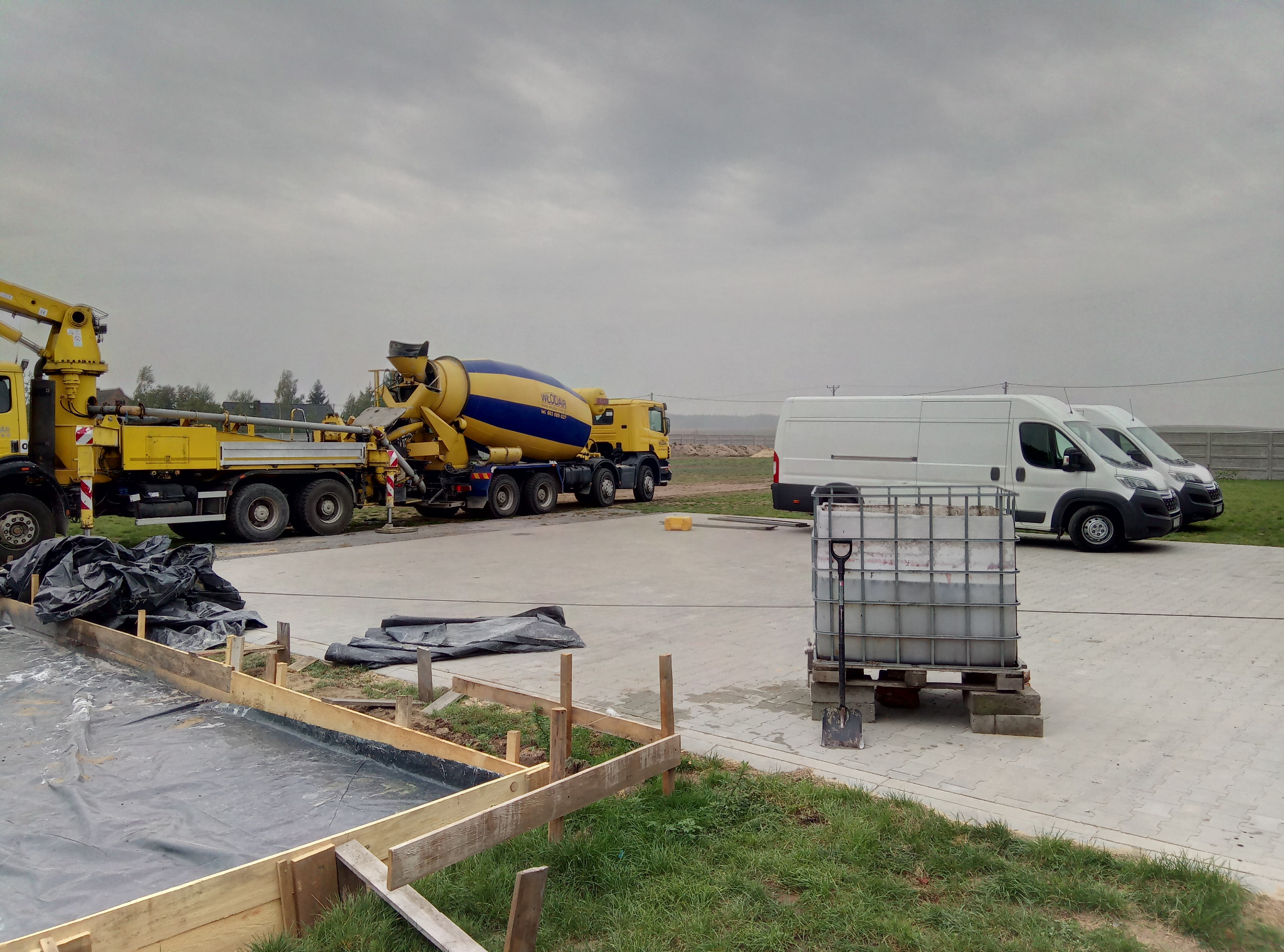 Przygotowanie pod wylewkę: szalunek z desek, folia izolacyjna, obok betoniarka i dwa busy na placu budowy w pochmurny dzień.