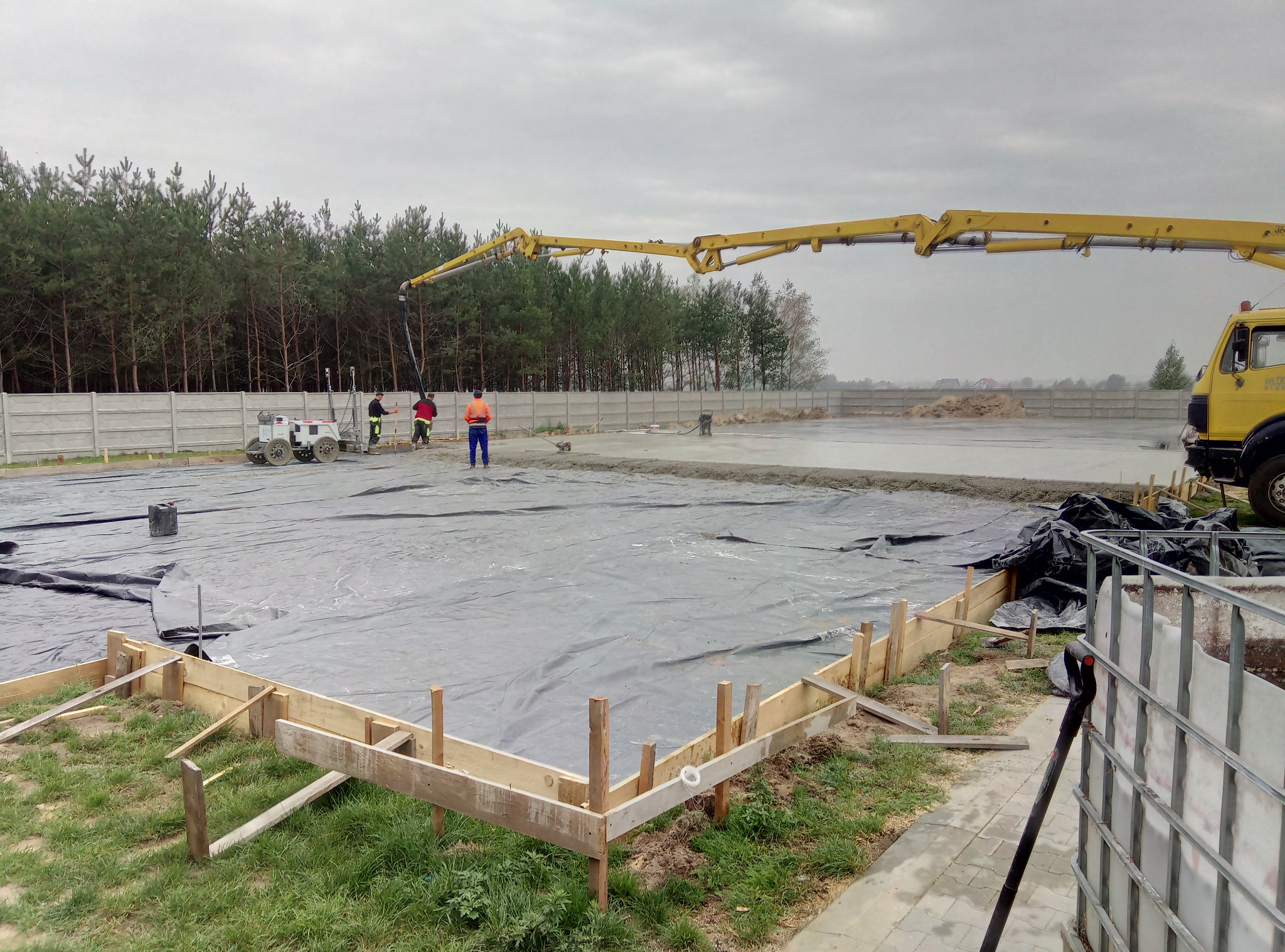 Wylewanie betonu na przygotowany teren z szalunkiem i folią, widoczny wysięgnik pompy do betonu oraz pracownicy w odblaskowych ubraniach.