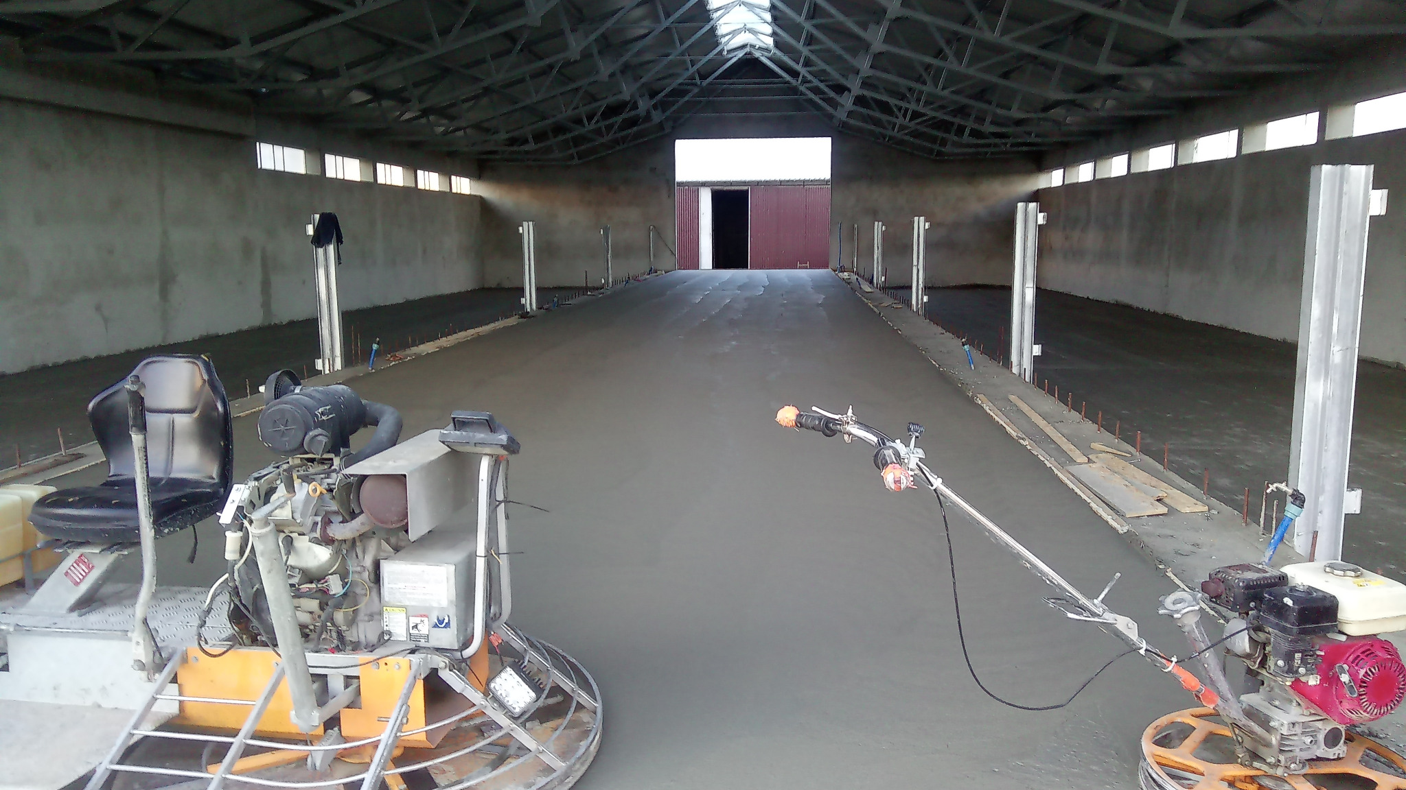 Wyrównywanie świeżo wylanej, szarej posadzki wewnątrz hali przemysłowej z widocznymi maszynami do zacierania i metalowymi słupami podtrzymującymi konstrukcję dachu.