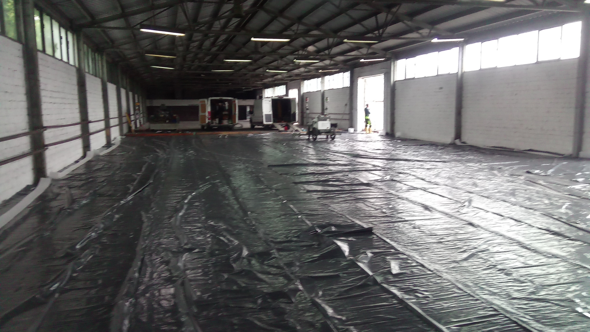 Przygotowanie podłoża pod wylewkę: rozłożona folia ochronna na dużej powierzchni hali przemysłowej, widoczne pojazdy i pracownik w oddali.