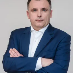 Portret biznesowy mężczyzny w niebieskim garniturze na neutralnym tle w Warszawie, z rękami skrzyżowanymi na piersiach i poważnym wyrazem twarzy.