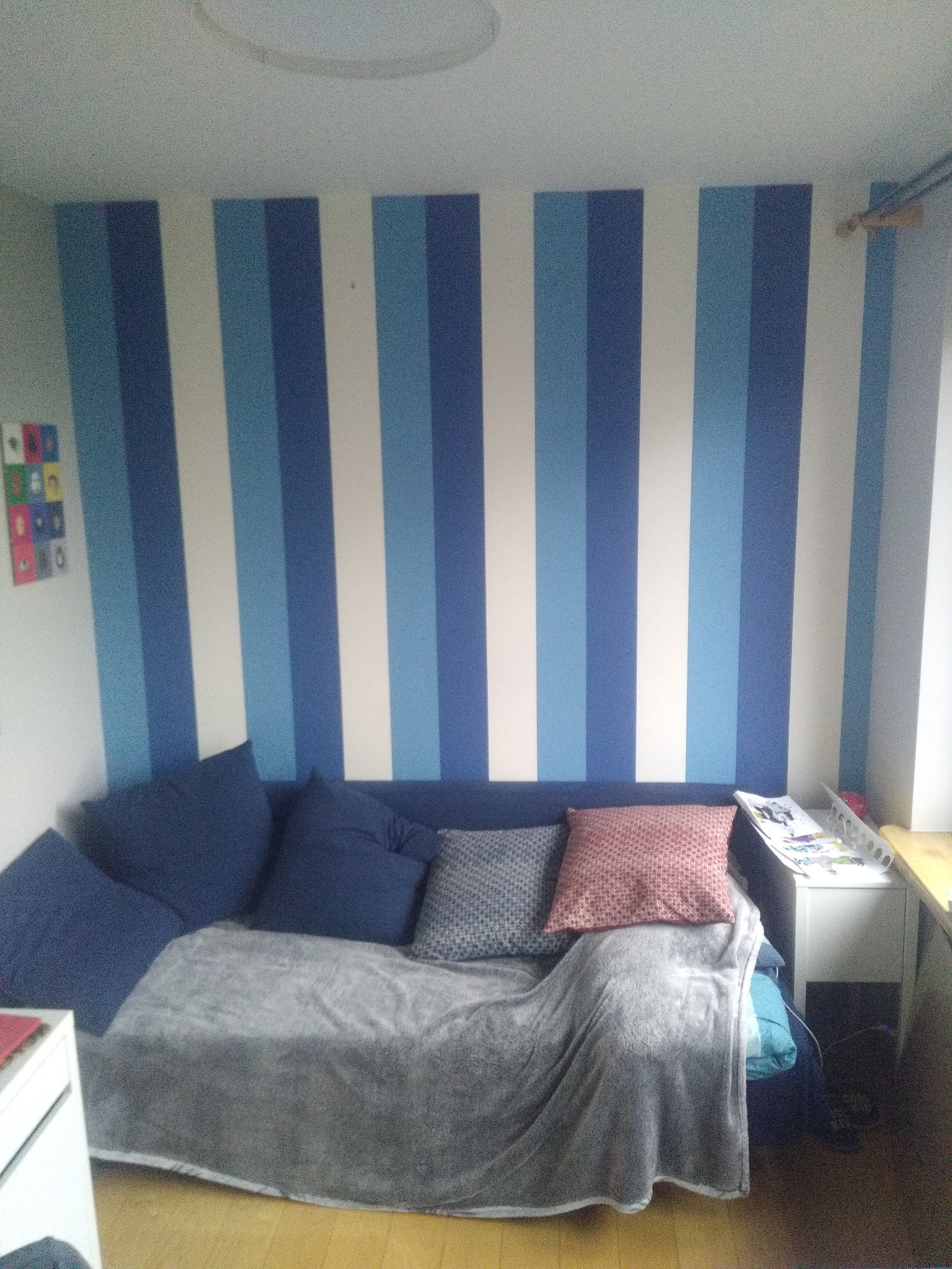 Pokój dziecięcy z łóżkiem/sofą, ściana w pionowe pasy w odcieniach błękitu i bieli, szare nakrycie i poduszki, biała szafka nocna przy oknie.