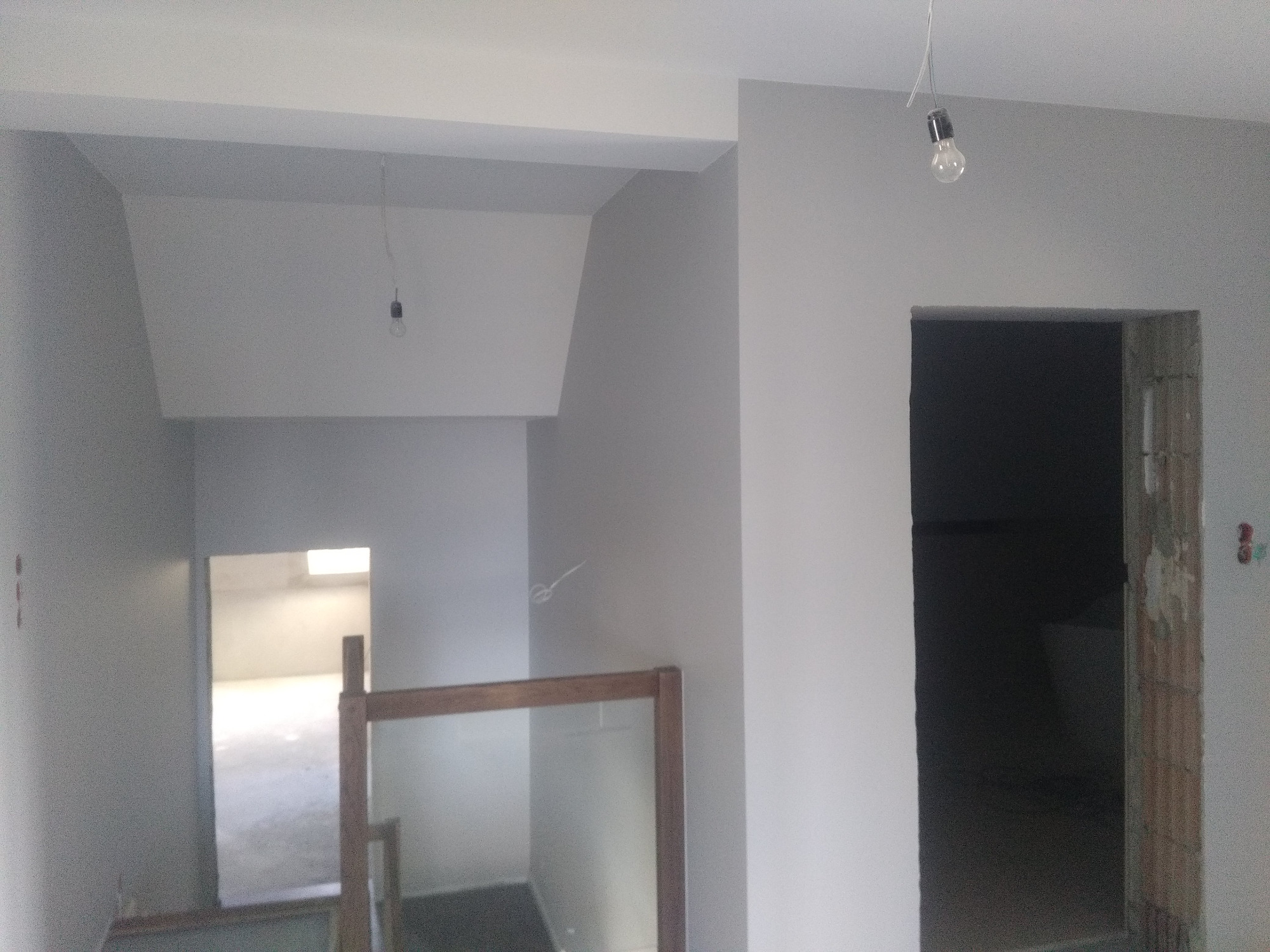 Wnętrze klatki schodowej w trakcie malowania na szaro, widoczne surowe żarówki na kablach, drewniana barierka schodowa i częściowo otynkowane wejście do pomieszczenia.