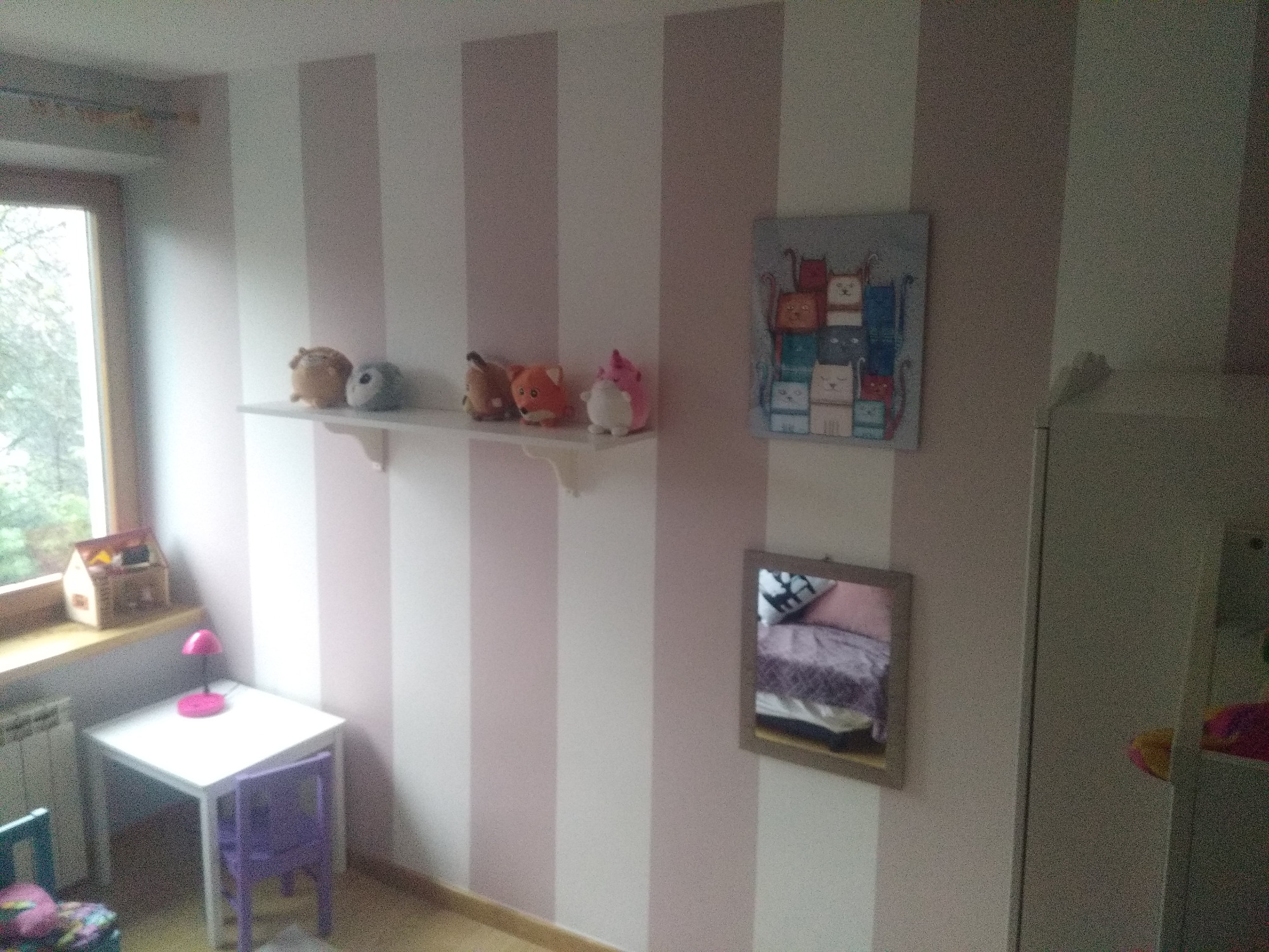 Pokój dziecięcy z pomalowanymi w pionowe pasy ścianami, biały stolik z fioletowym krzesełkiem, półka z pluszakami i lustro odbijające fragment łóżka.