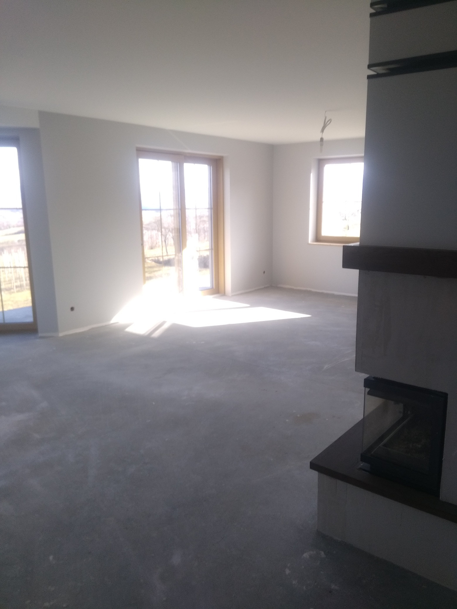 Surowe wnętrze z wylewką betonową, widoczne okna i fragment kominka z szybą, przygotowane do dalszych prac wykończeniowych.