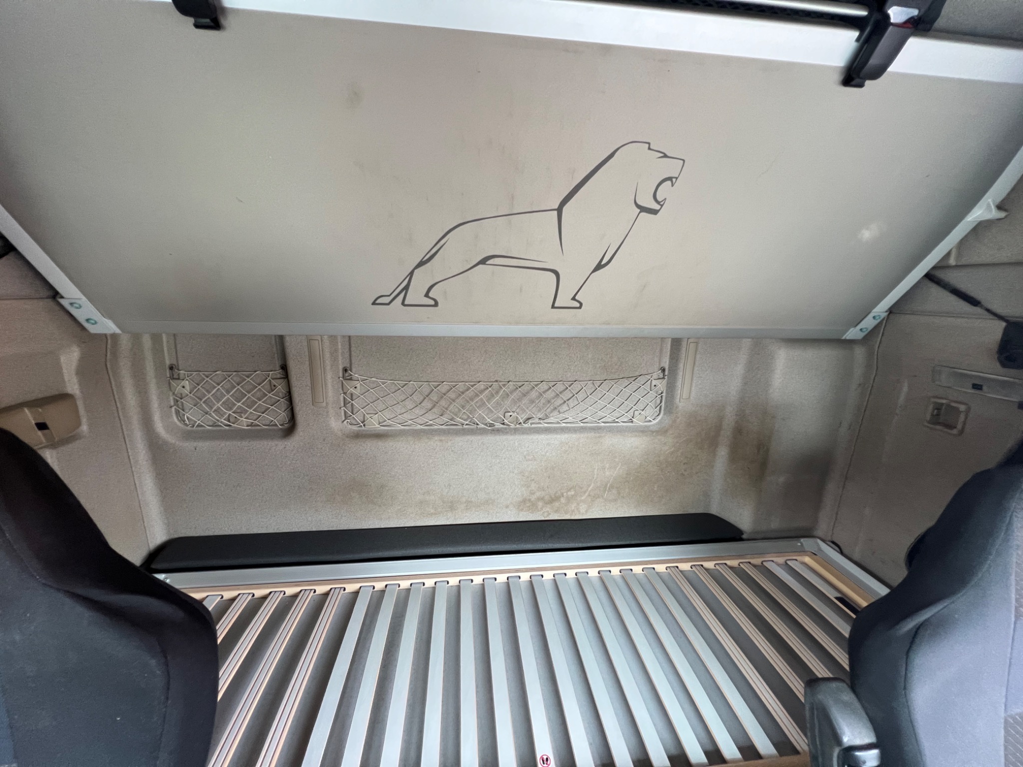 Wnętrze kabiny ciężarówki z rozłożonym łóżkiem, widoczna siatka bagażowa i grafika lwa na suficie.