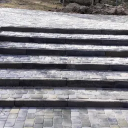 Schody z kostki brukowej prowadzące do podwyższonego tarasu, z widocznymi elementami drewnianej konstrukcji i fragmentem ogrodu.