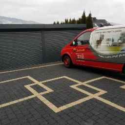 Szara brama garażowa z lamelami, obok zaparkowany czerwony samochód dostawczy z reklamą firmy produkującej systemy rolet, bram i moskitier, na pierwszym planie kostka brukowa z jasnymi liniami.