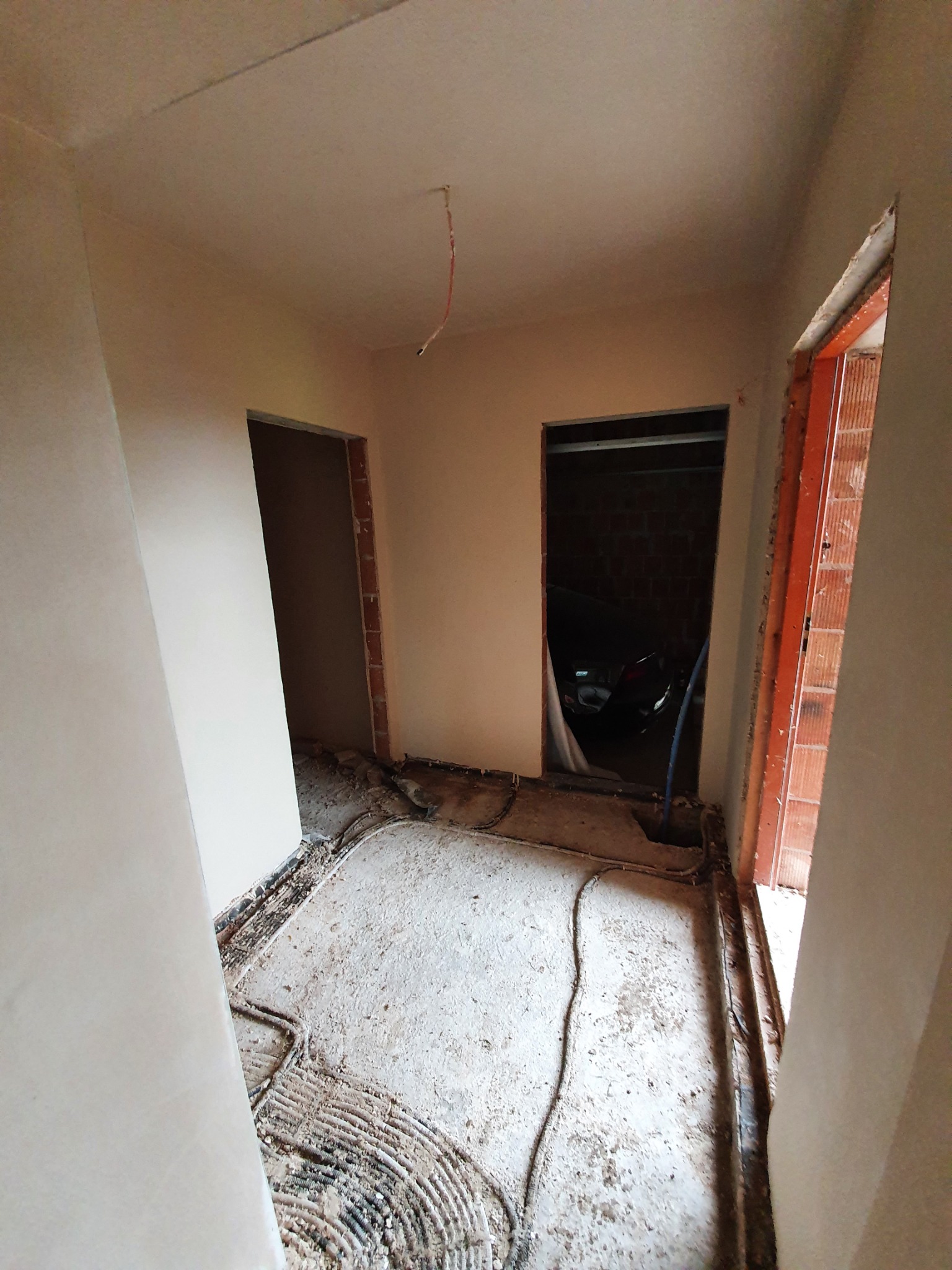 Świeżo otynkowane ściany wewnętrzne w trakcie prac wykończeniowych, widoczne przewody na podłodze i fragment samochodu w garażu przez otwarte drzwi.