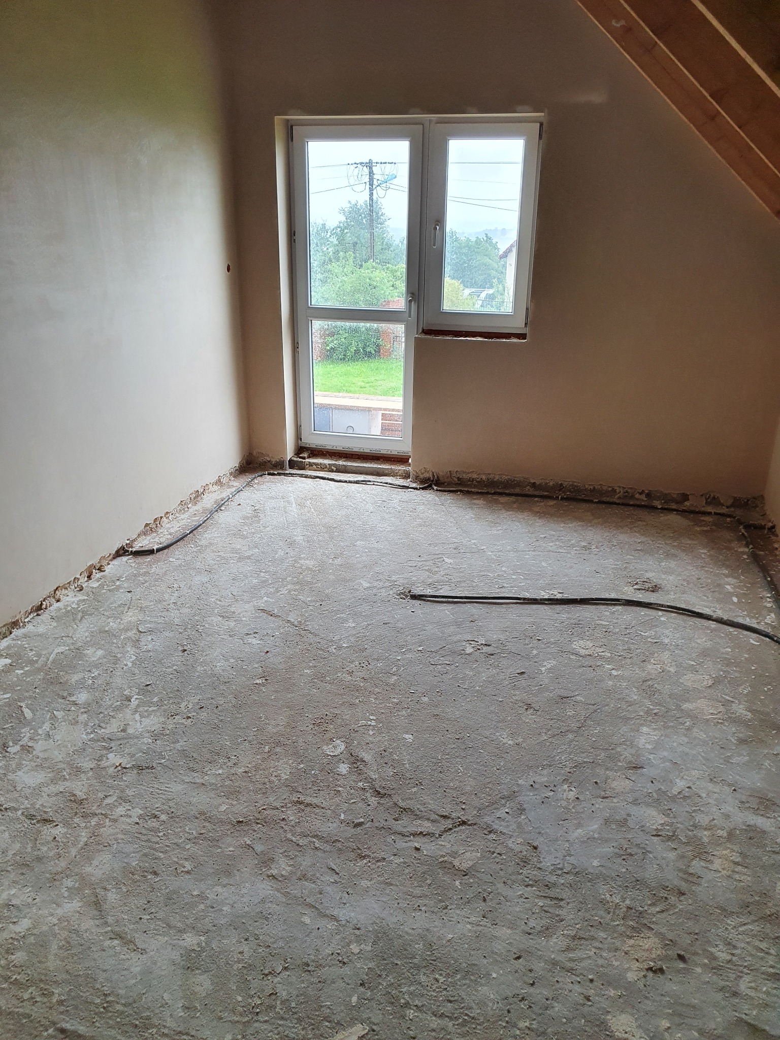 Pokój na poddaszu w trakcie prac wykończeniowych, ściany po nałożeniu gładzi gipsowej, widoczne przewody elektryczne przy podłodze, surowe drewniane elementy konstrukcji dachu.