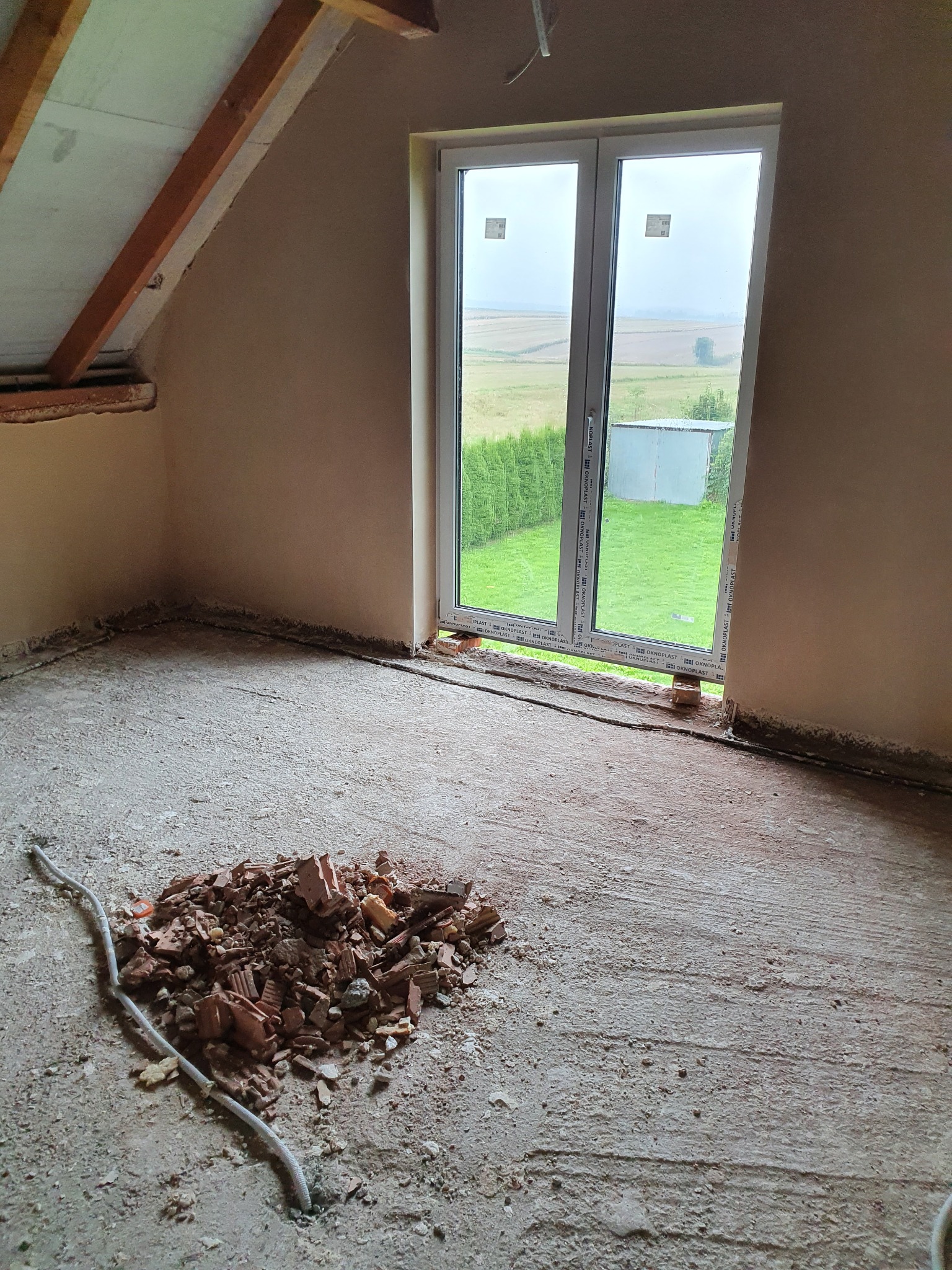 Pokój w Igołomi po nałożeniu tynków, widoczne surowe wykończenie, ściany otynkowane, gruz na podłodze, okno z widokiem na zewnątrz.