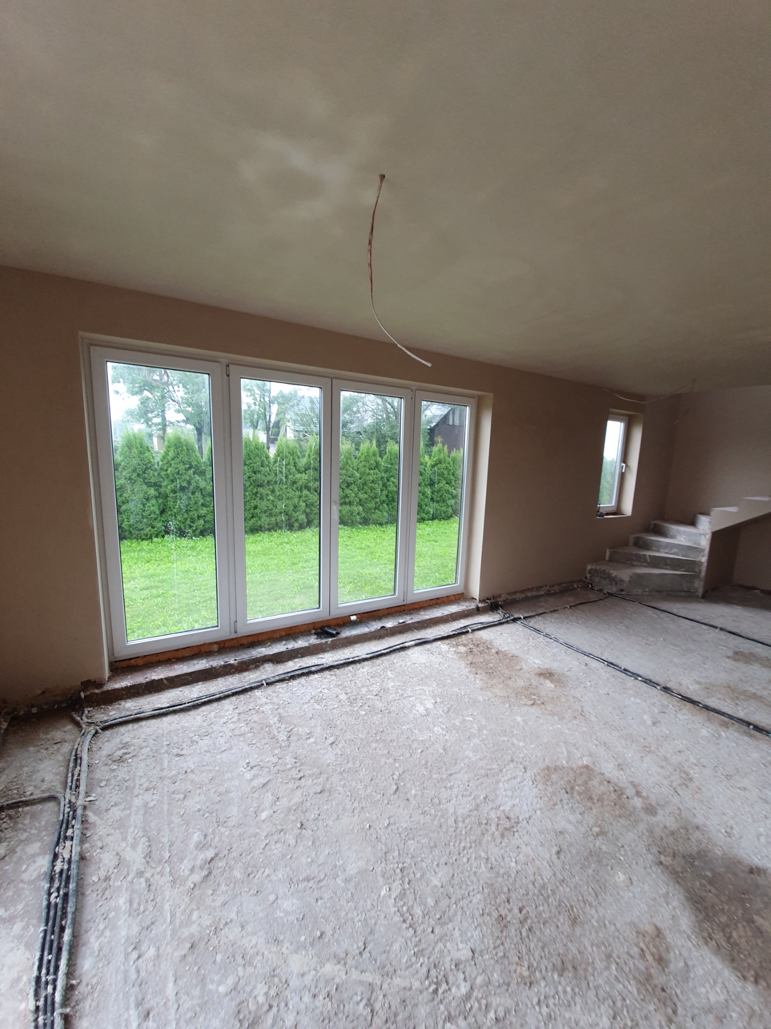 Wnętrze w stanie deweloperskim po nałożeniu świeżych, beżowych tynków na ścianach i suficie, z widocznymi przewodami elektrycznymi, oknami z widokiem na zieleń i surowymi schodami.