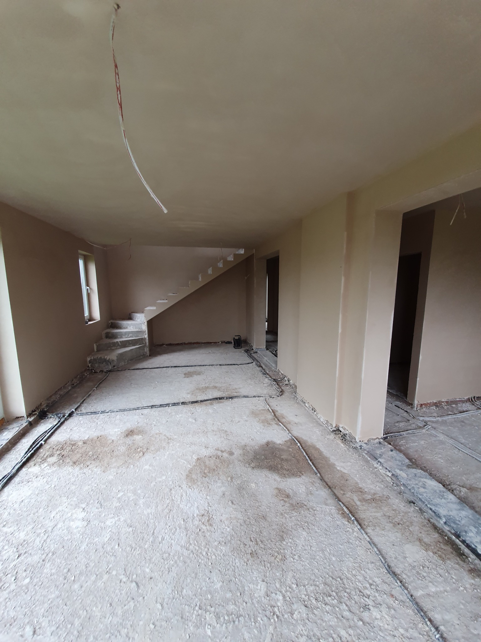 Wnętrze domu w trakcie prac wykończeniowych, ściany i schody pokryte świeżym, jasnym tynkiem, widoczne kable instalacji elektrycznej na podłodze.