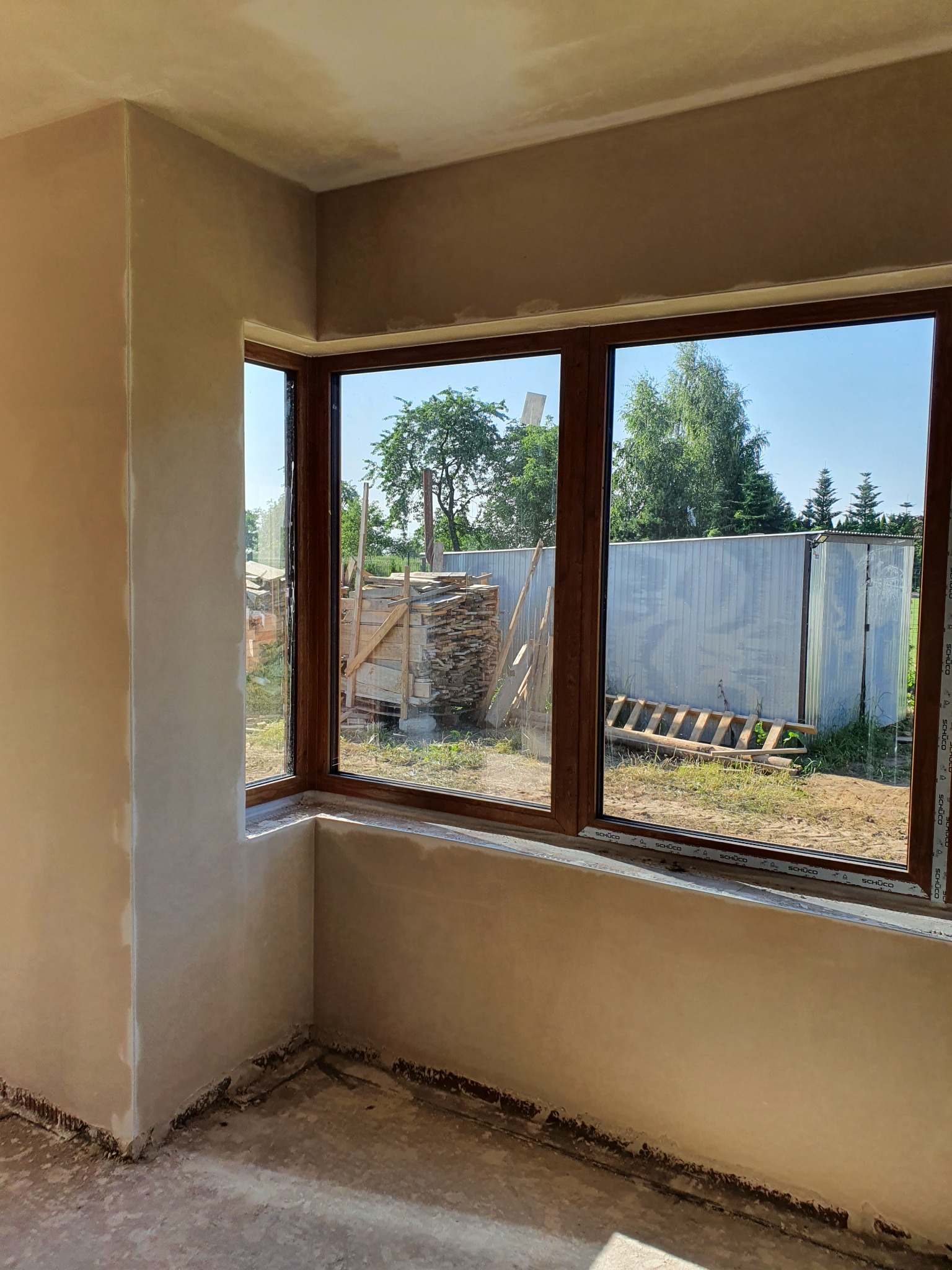 Wnętrze pokoju z świeżo nałożonym tynkiem na ścianach, widoczne trójdzielne okno z drewnianą ramą i widokiem na podwórko z stertą drewna i metalową szopą.